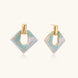 Geometric square enamel earrings with marbled turquoise and lavender pattern, gold U-shaped connector on cream background.