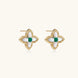 Gold quatrefoil stud earrings with white mother-of-pearl petals and emerald green gemstone center on beige background.