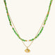 Layered necklace with green stone beads, gold spacers, white accents, and protective eye pendant on cream background.
